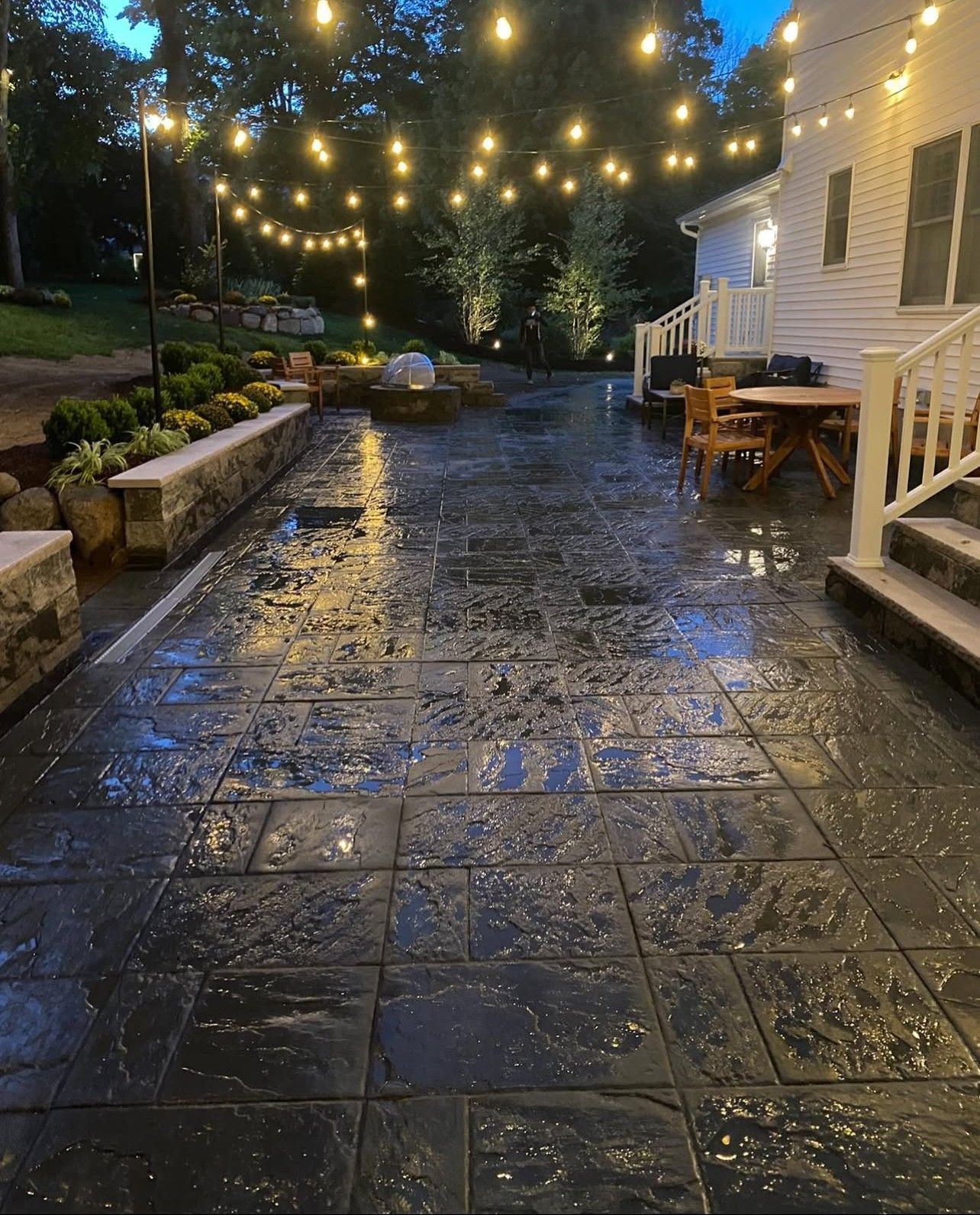 Patio with wet, dark stamped concrete, string lights, and a white house at dusk.
