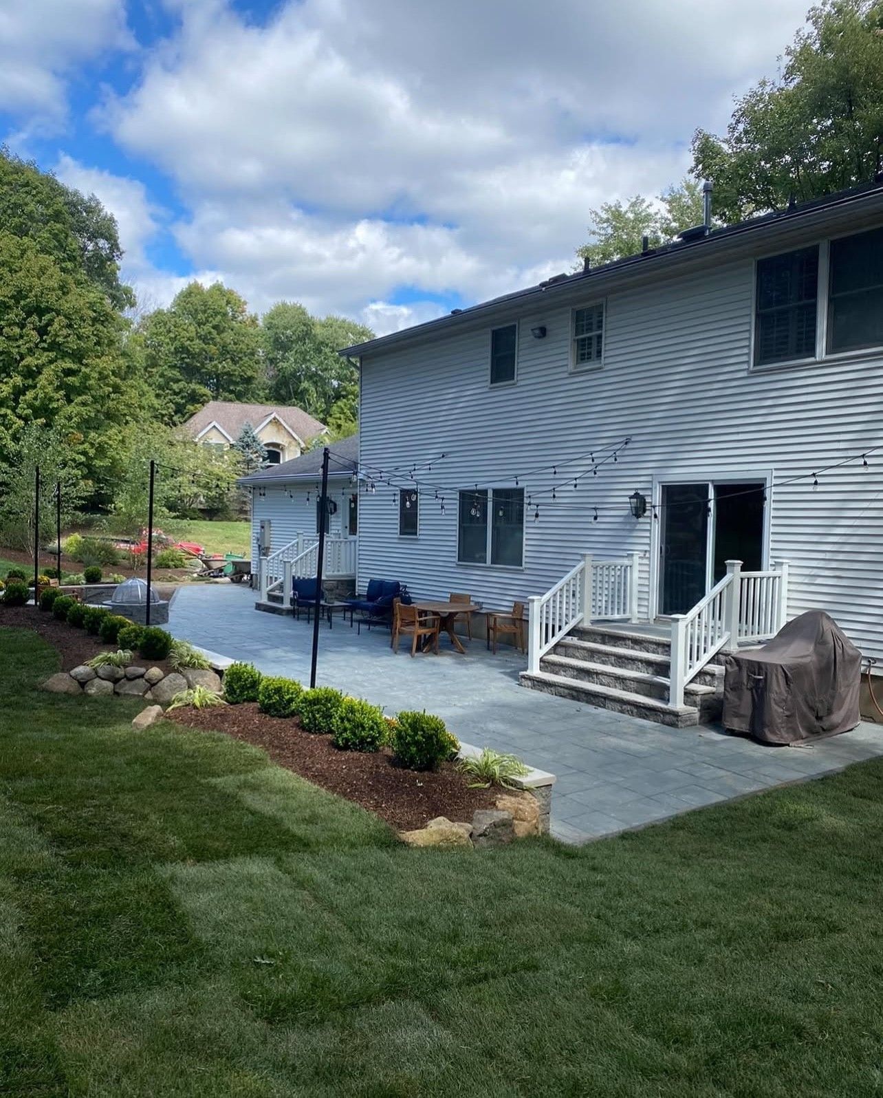 Backyard with patio, landscaping, two-story house. Cloudy sky, green grass, stone retaining wall, and string lights.