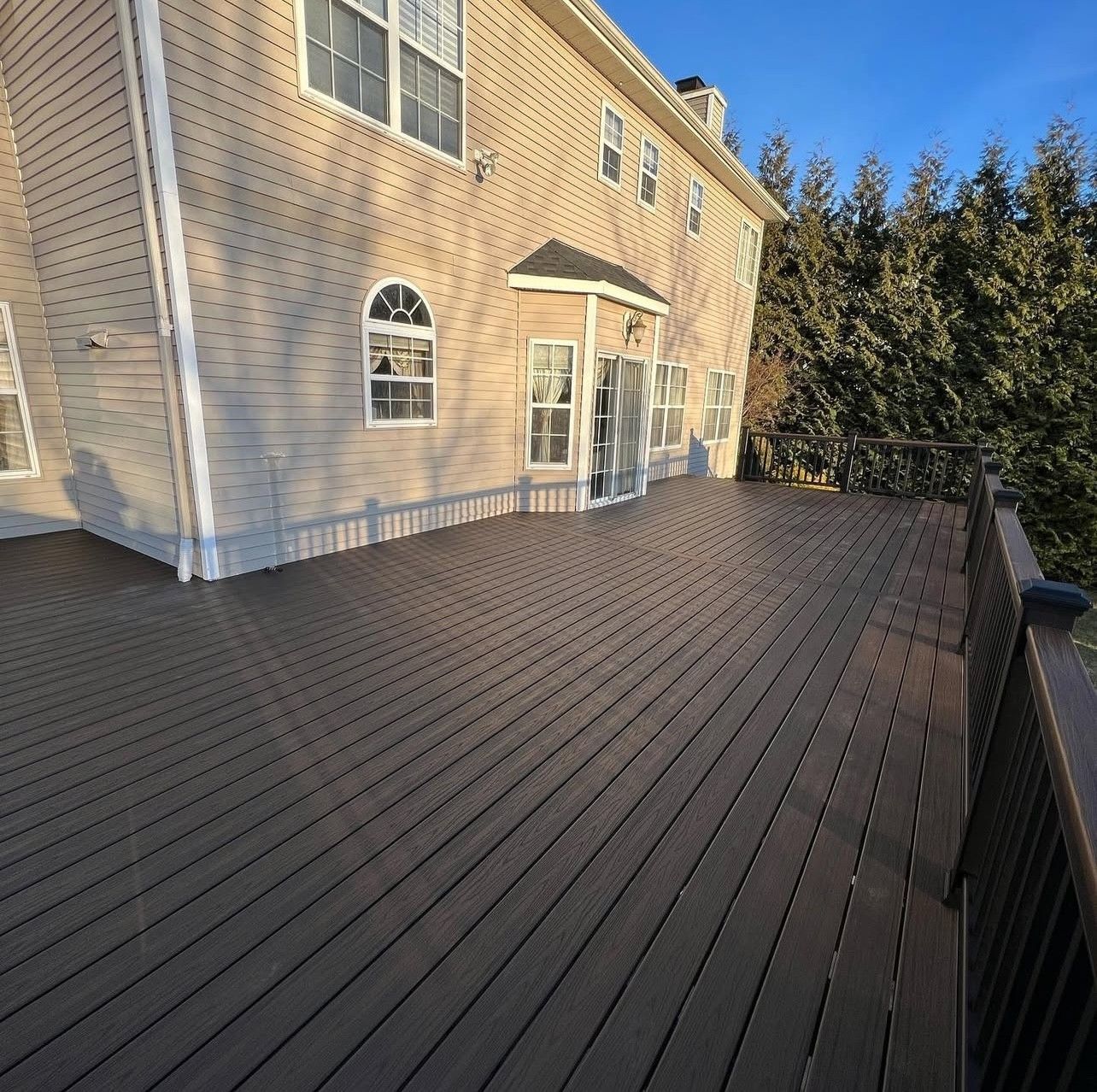 Composite deck outside a two-story house. Brown deck boards, beige siding, and dark railing.