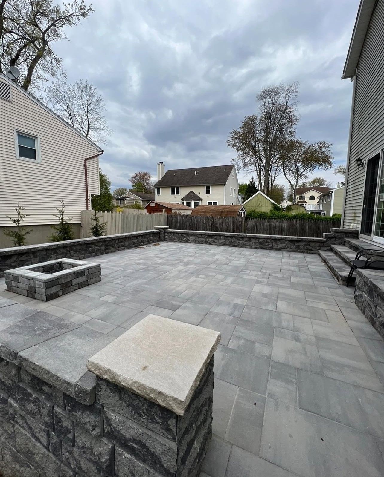 Gray paver patio with a fire pit, retaining walls, and houses in the background under a cloudy sky.