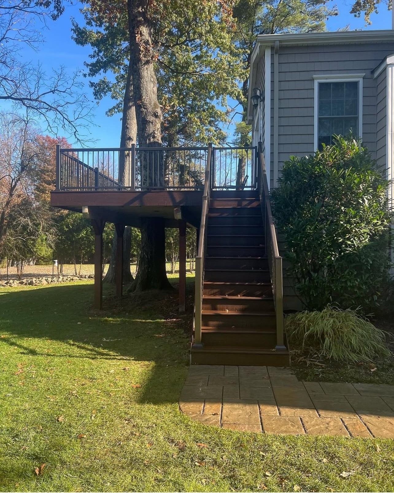 A wooden deck built around a tree with stairs, next to a gray house with green grass and a blue sky.