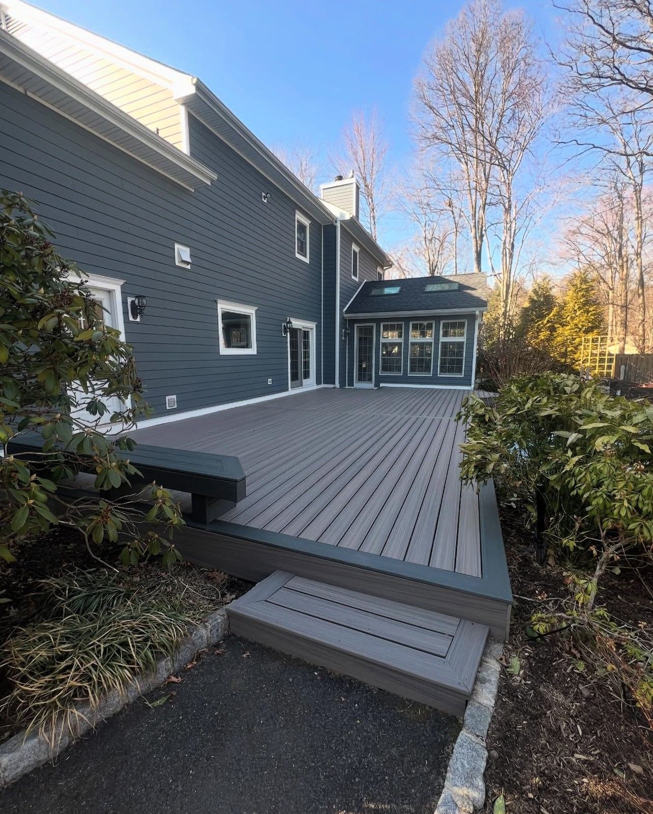 Dark gray composite deck attached to a blue house with a small sunroom; landscaping surrounds.