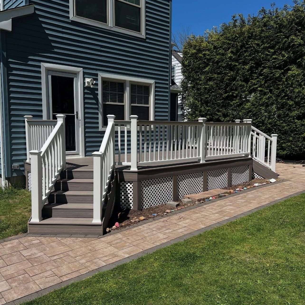 Blue house with white deck and stairs, brown brick path, green grass, and a shrub.