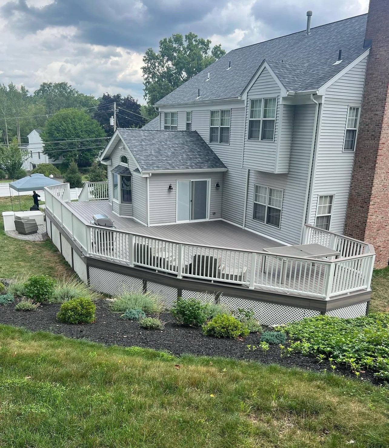 Two-story house with a gray deck and white railings. Green landscaping and dark mulch border the deck.