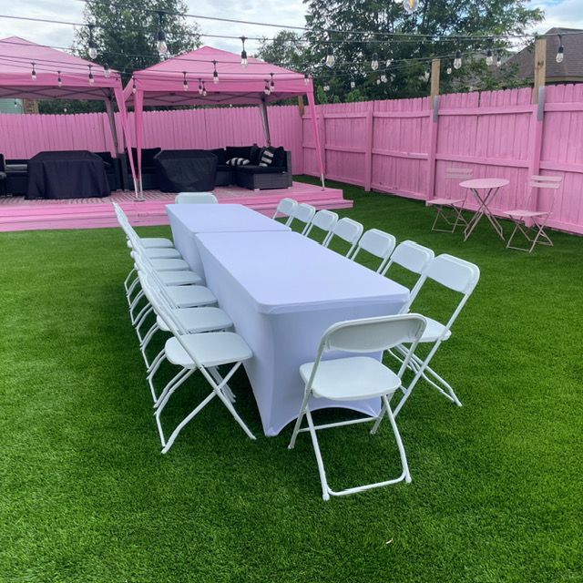 A long table with a white tablecloth and folding chairs set up on a lawn in front of a pink wooden fence and canopy tents.