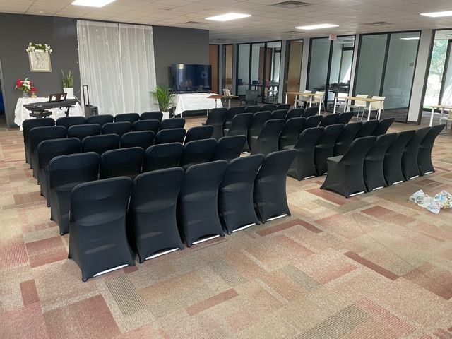 Rows of black-covered chairs arranged in a meeting room with carpeted floors, white curtains, and a television screen.