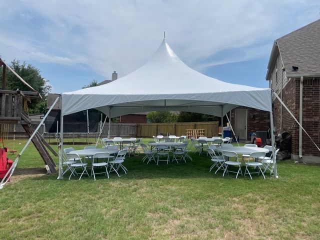 A white high-peak party tent set up in a residential backyard, featuring several round tables with chairs.