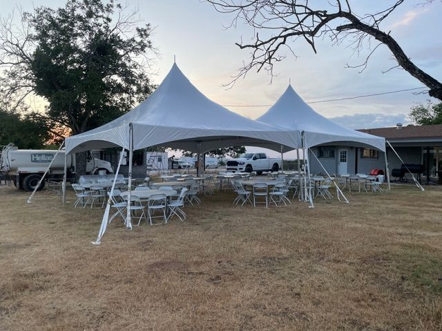 Two large, white, high-peak event tents set up on a grassy field with round tables and chairs underneath.