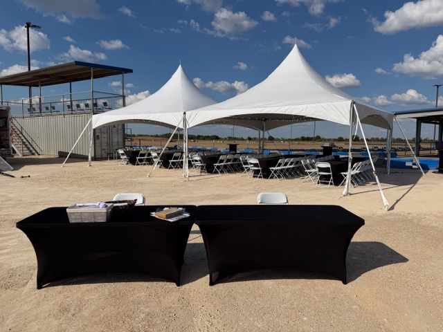 Two large white tents set up for an outdoor event with black-clothed tables and chairs on a sandy ground under a blue sky.