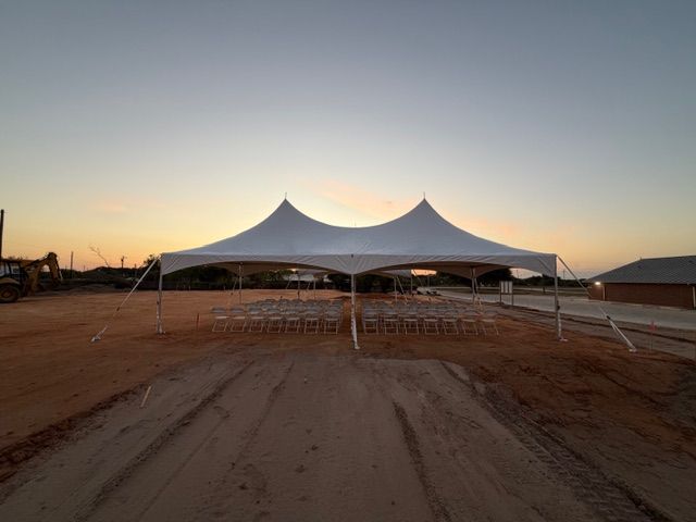 A white frame tent set up on a dirt lot at dusk, with rows of chairs visible underneath.