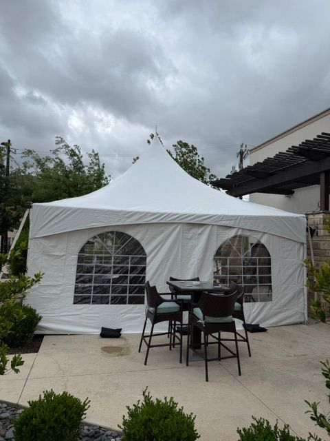 A white high-peak party tent with arched window walls stands on a patio with a small bistro table and chairs nearby.
