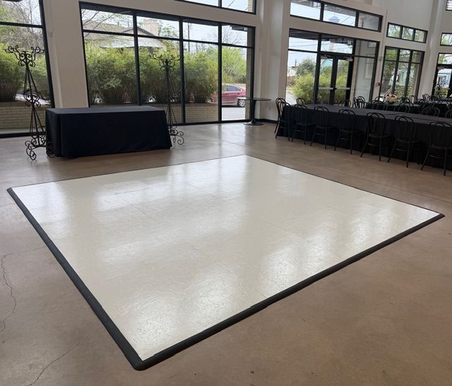 A white, square portable dance floor with black trim centered in a room with large windows and tables with black cloths.
