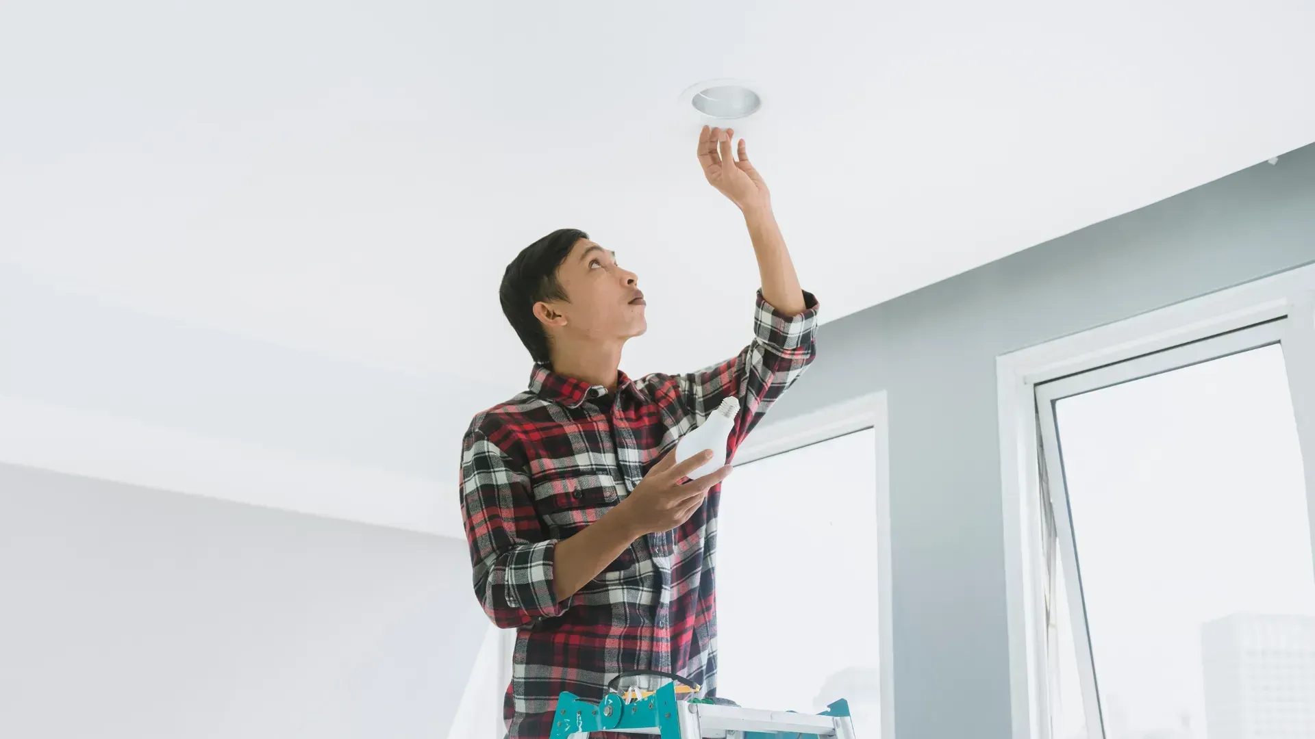A man is standing on a ladder fixing a light fixture.