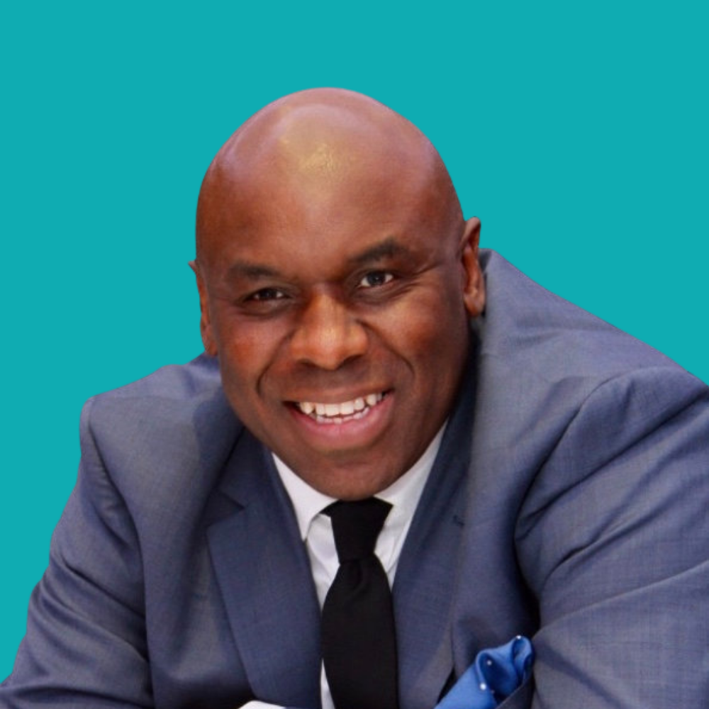 David Shosanya in a grey suit, smiles, leaning forward against a turquoise background. He wears a black tie and has a blue pocket square.