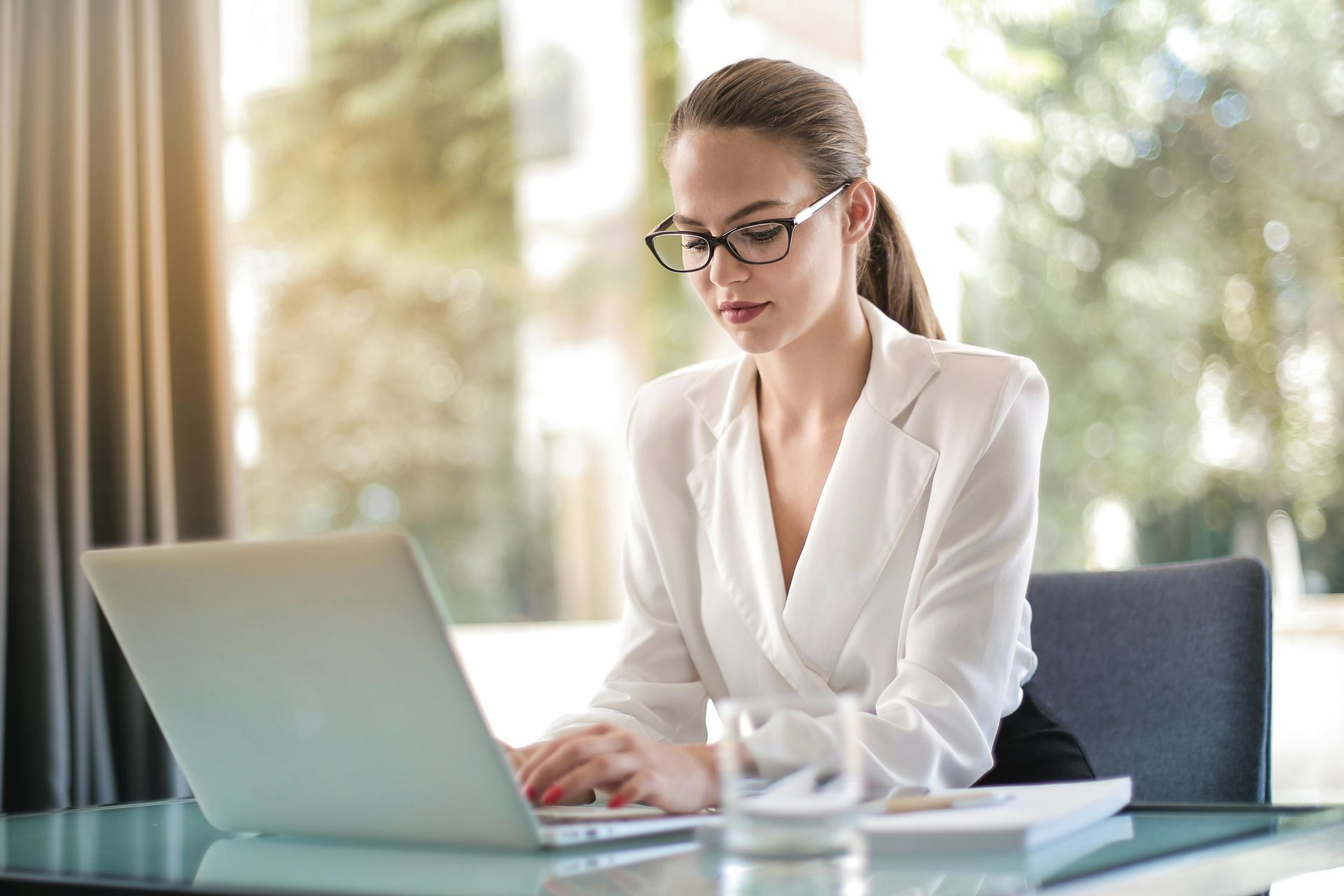 Woman in white blazer working on laptop at a desk, wearing glasses, in an office setting.