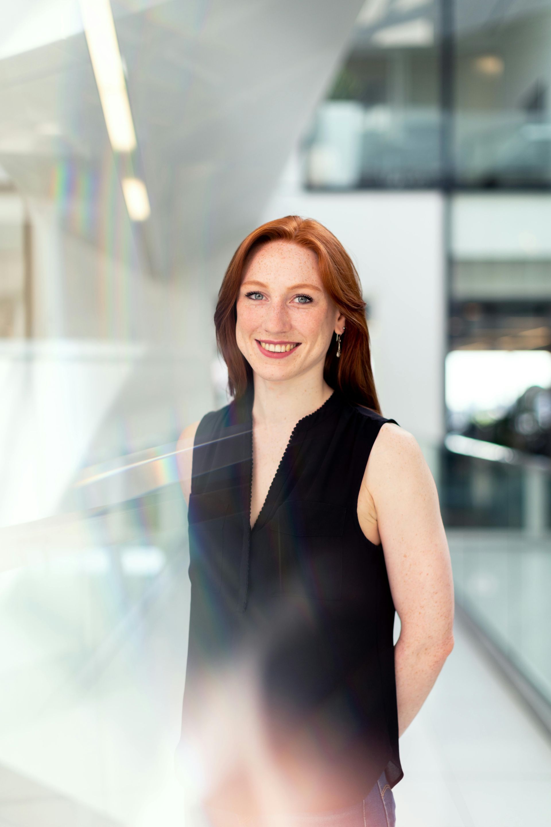 Woman with red hair, smiling, in black sleeveless top, standing in a modern building hallway.