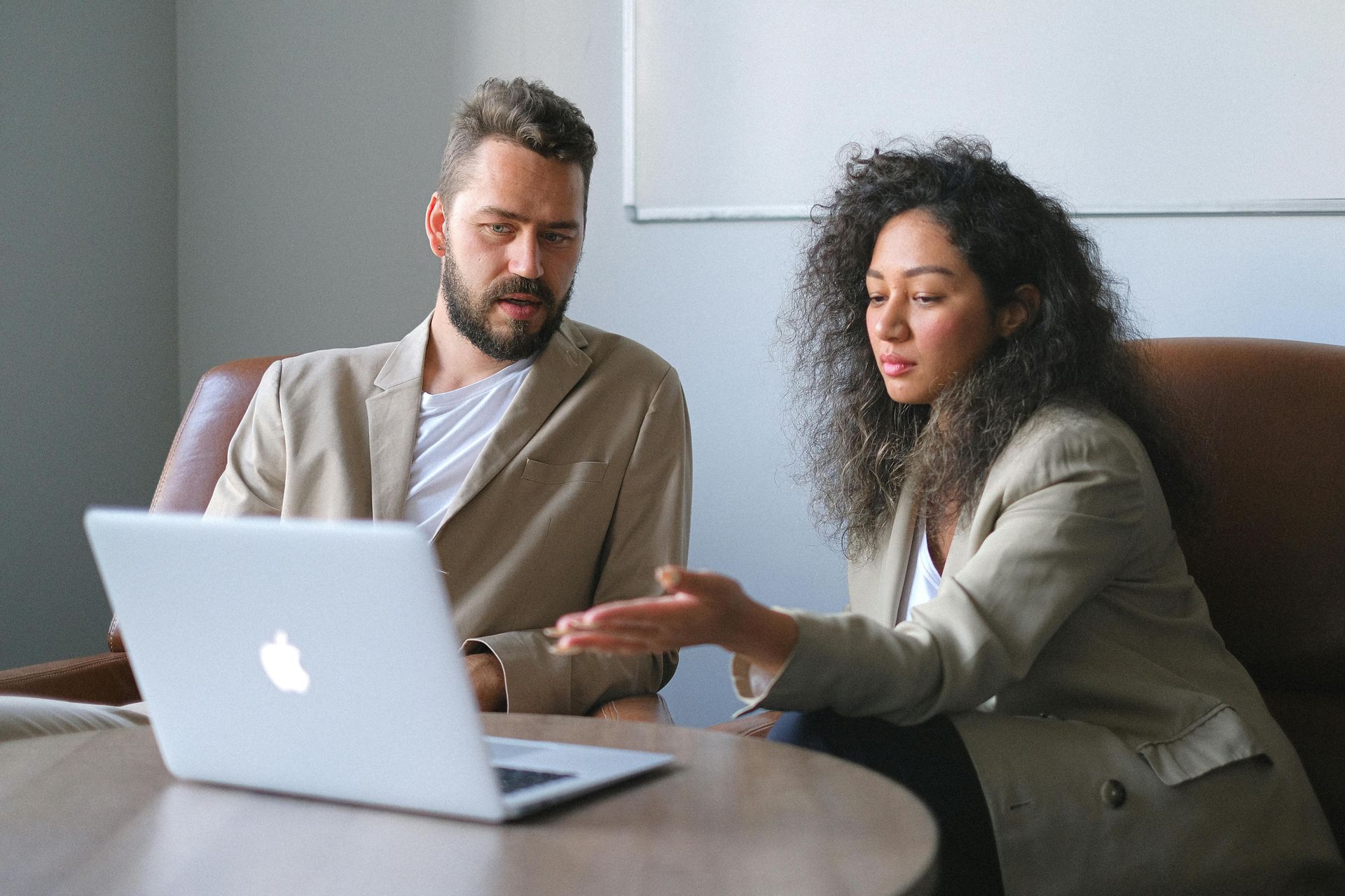 Man and woman looking at a laptop, discussing something. Woman points to screen, both are in suits.