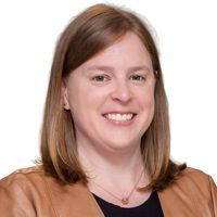 Woman in a tan jacket smiles, facing forward. Light brown hair, black top, simple necklace, white background.