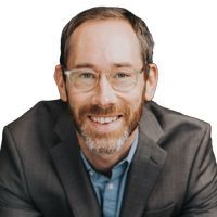 Man with a beard and glasses smiling, wearing a gray suit and a blue button-down shirt.
