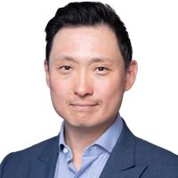 Man in a blue suit and light blue shirt smiles at the camera against a white background.