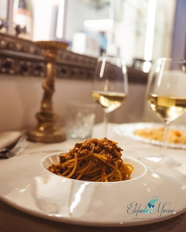 Un piatto di spaghetti al ragù è appoggiato su un tavolo accanto a due bicchieri di vino bianco