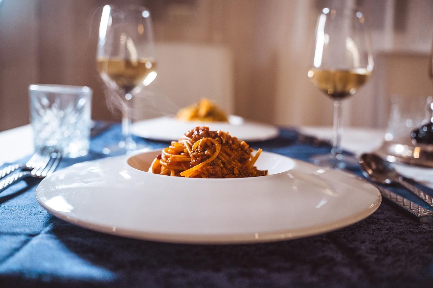 Un piatto bianco con una porzione di spaghetti su una tovaglia blu, con due calici di vino sullo sfondo.