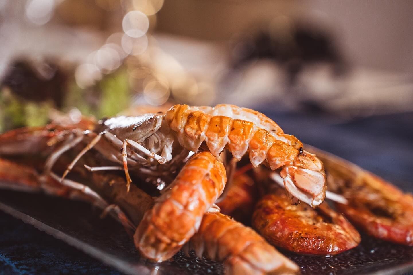 Primo piano di scampi grigliati serviti su un piatto rettangolare scuro, con una luce di sfondo morbida e sfocata.