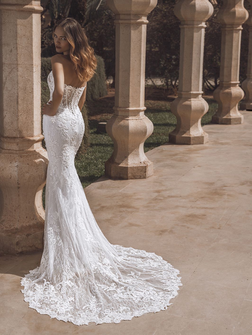 Frau in weißem Spitzenbrautkleid mit langer Schleppe posiert im Freien neben Steinsäulen.