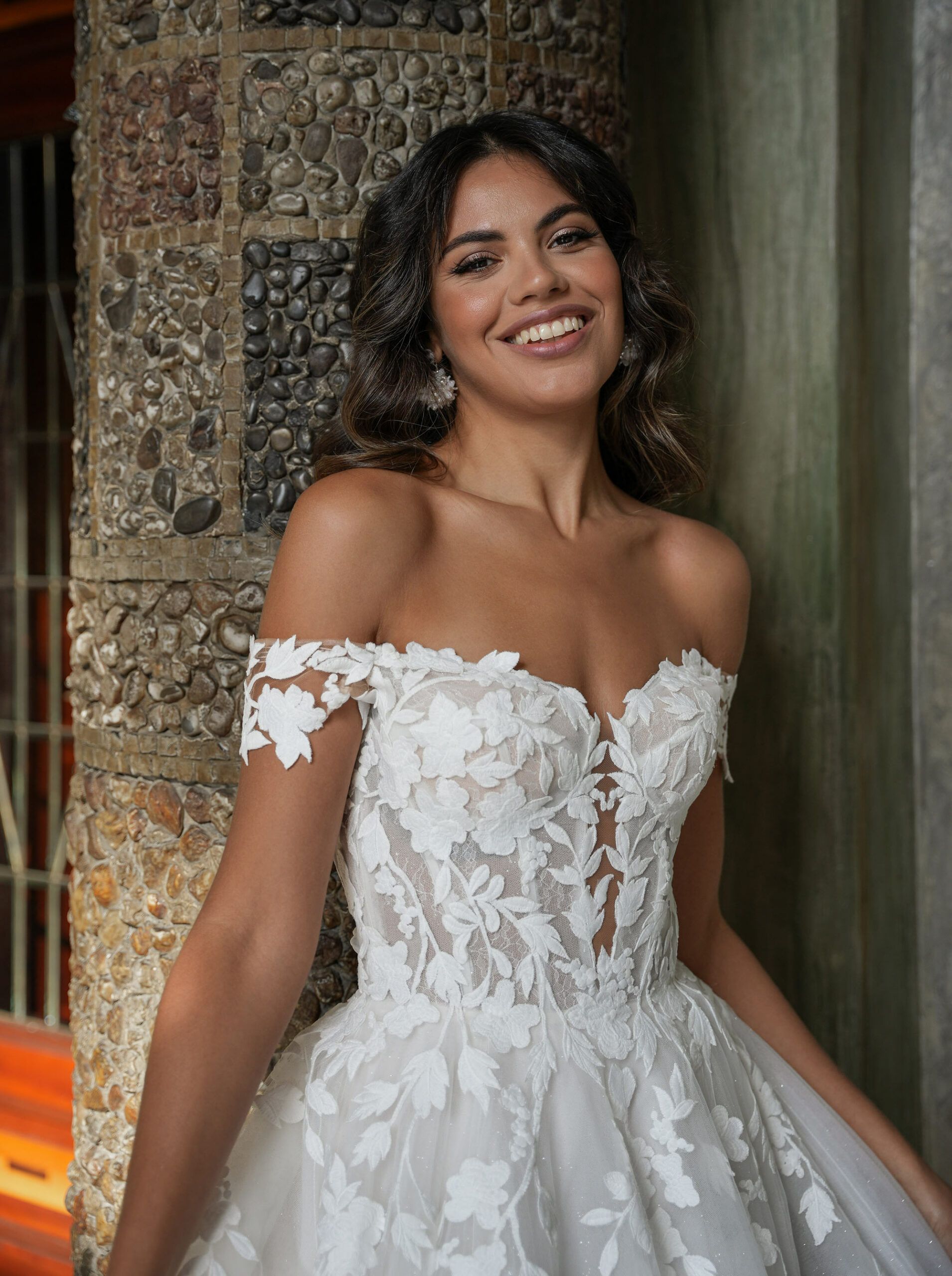 Eine Frau in einem Brautkleid steht neben einer Steinmauer und lächelt.