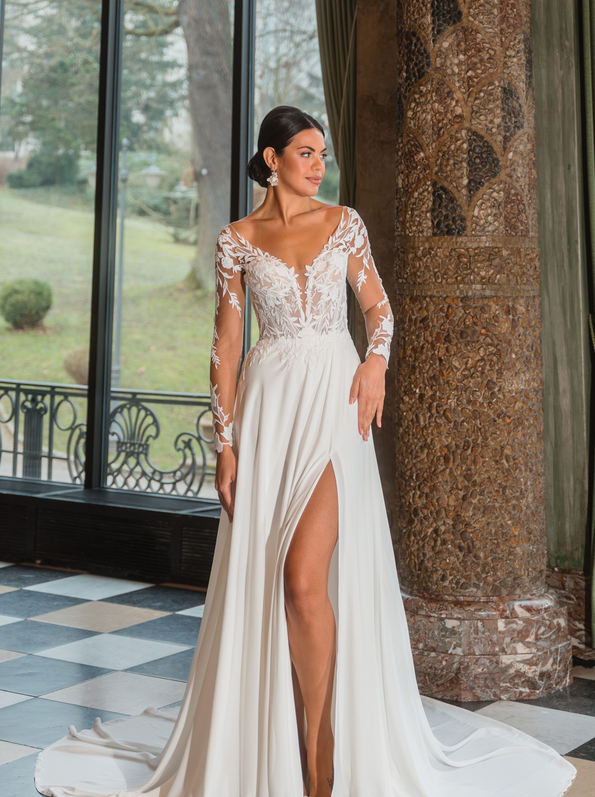Frau in weißem Brautkleid mit hohem Schlitz steht in einem Raum mit großen Fenstern und Marmorsäulen.