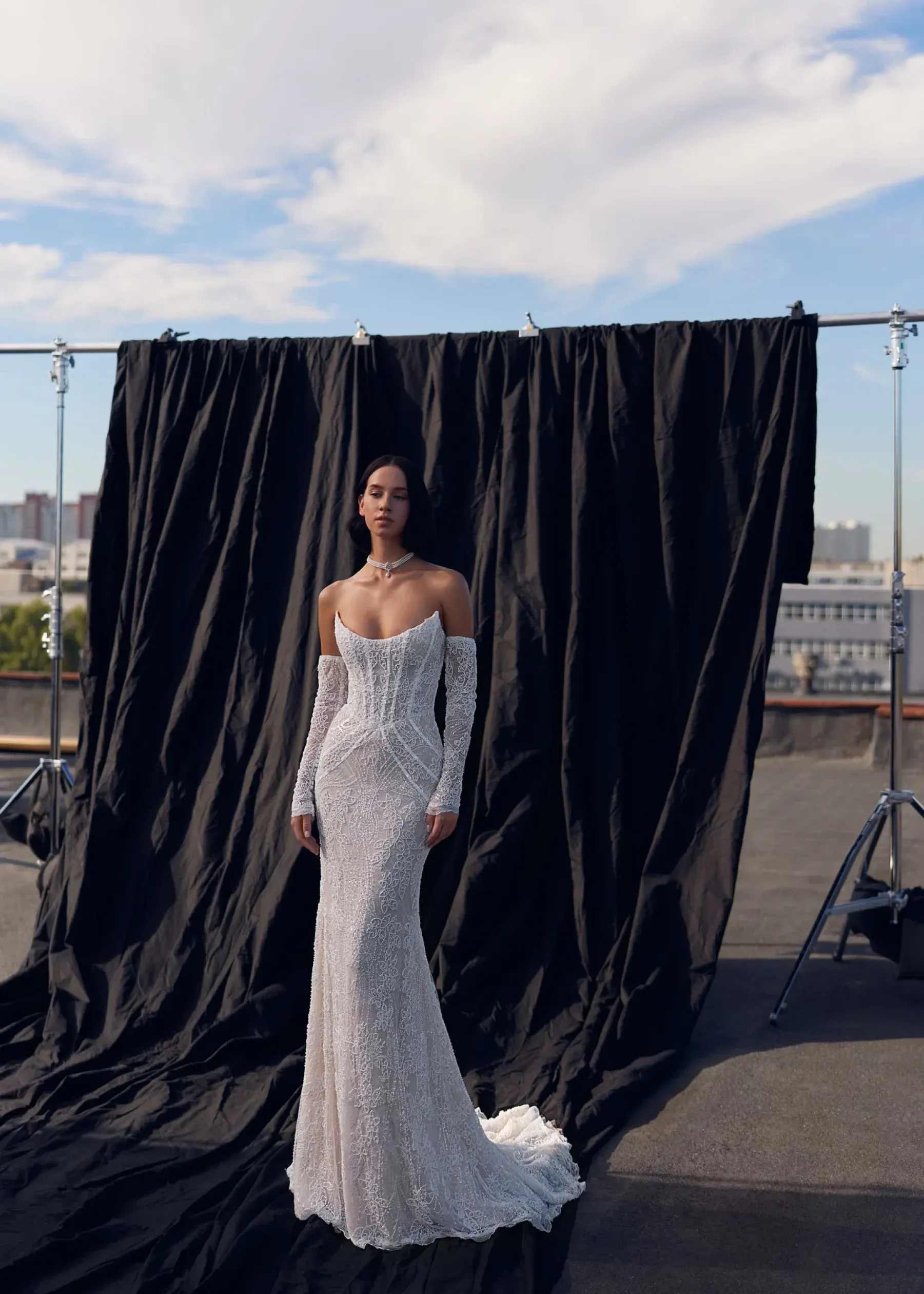 Frau in einem verzierten schulterfreien Kleid posiert auf einem Dach. Schwarzer Hintergrund und blauer Himmel.