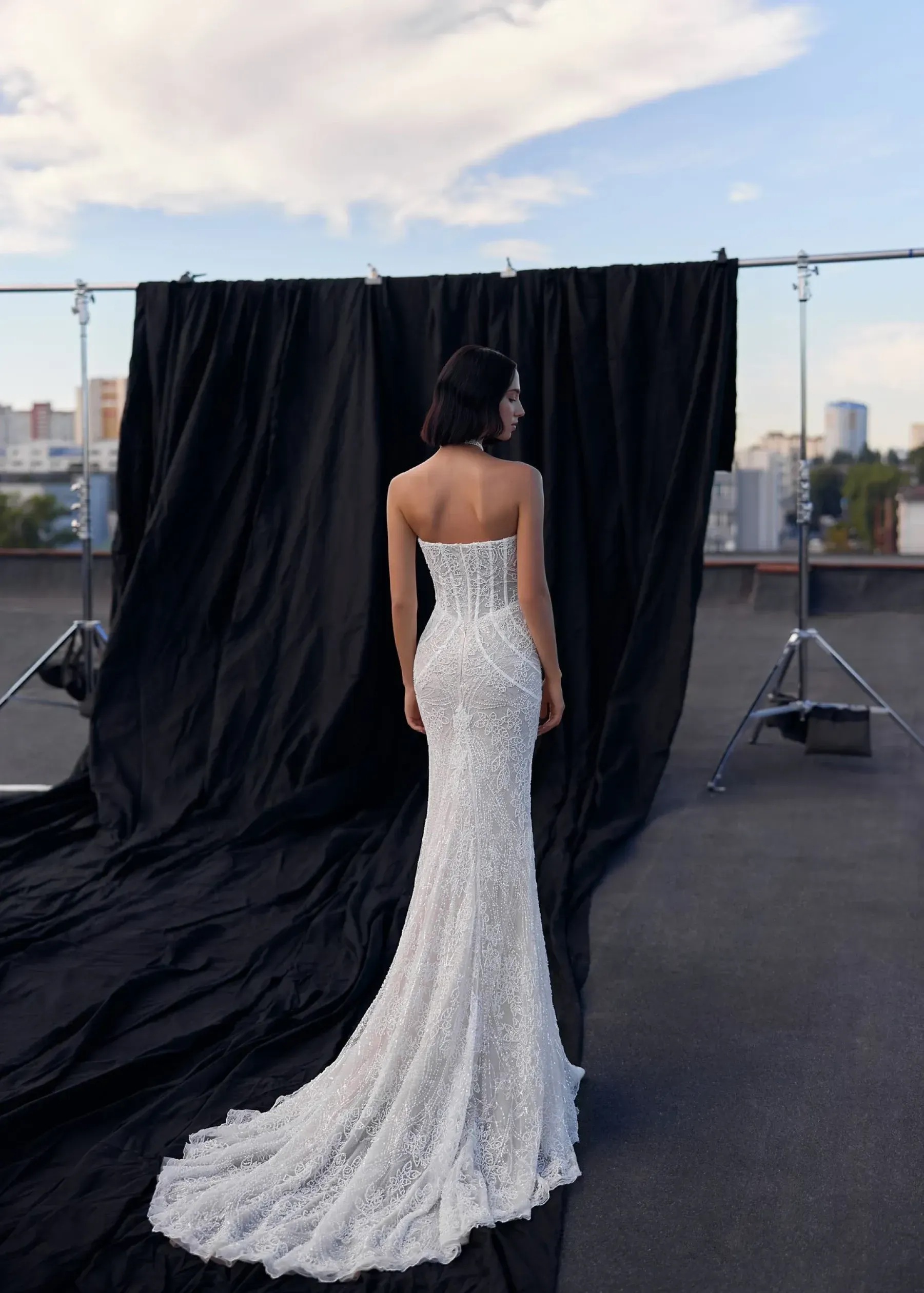Frau in einem funkelnden weißen Kleid mit langer Schleppe, die vor einem schwarzen Hintergrund auf einem Dach steht.
