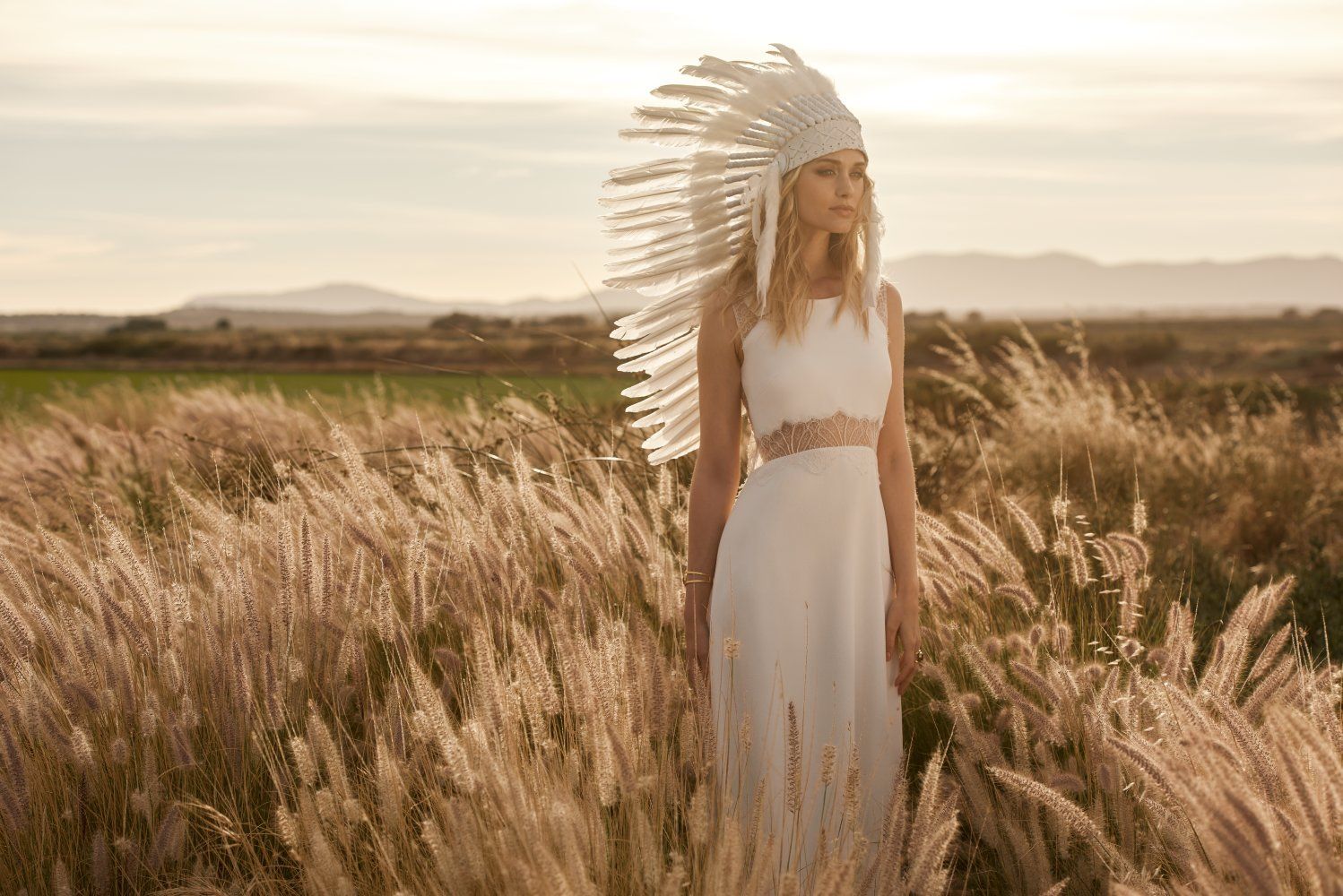 Eine Frau in einem weißen Kleid und einem indianischen Kopfschmuck steht auf einem Feld mit hohem Gras.