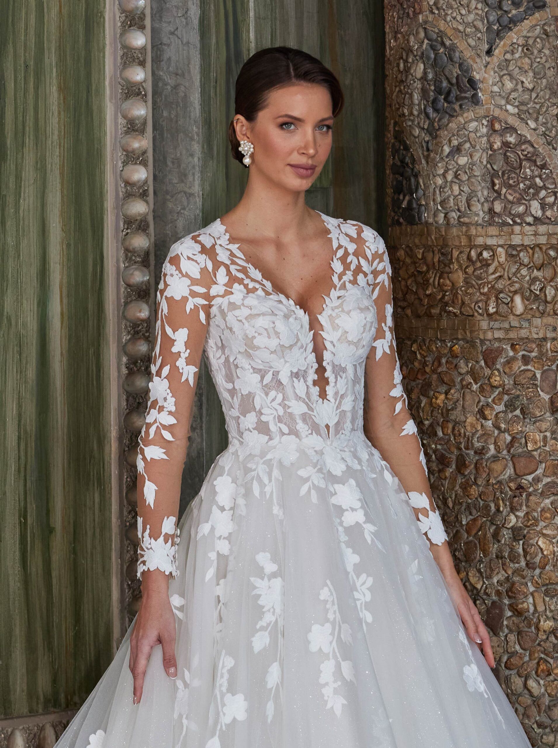 Frau im weißen Spitzenbrautkleid, V-Ausschnitt, lange Ärmel, lächelnd. Steht neben dekorierter Säule.