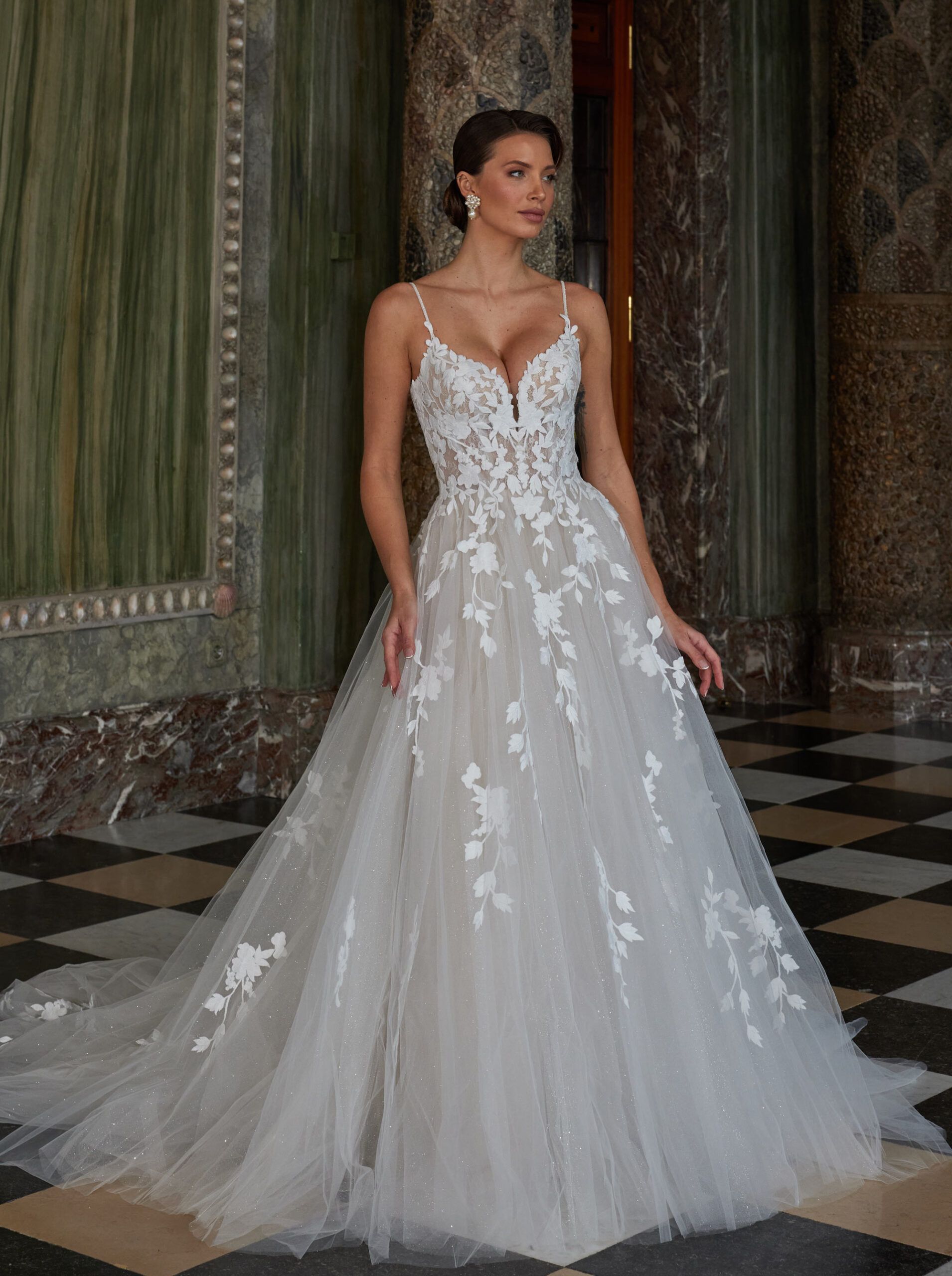 Frau in einem weißen geblümten Brautkleid mit Spaghettiträgern steht neben Marmorsäulen.