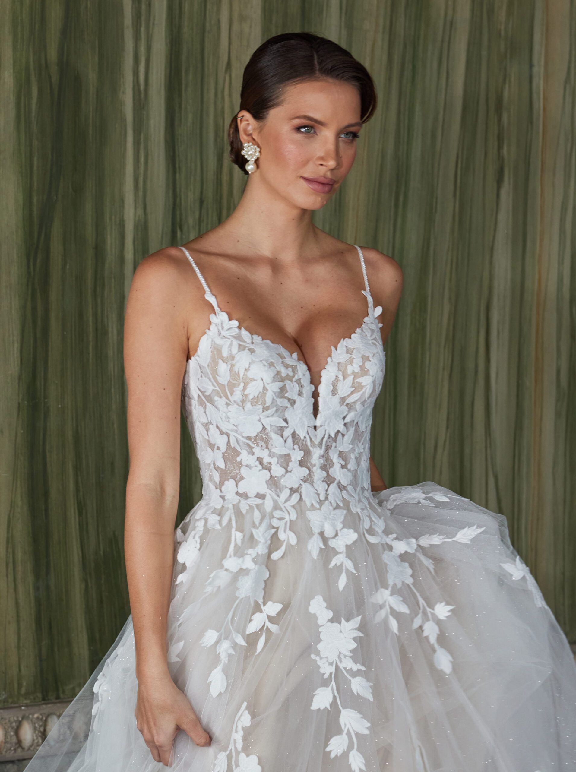 Frau in einem weißen Spitzenbrautkleid mit tiefem Ausschnitt, steht vor einem strukturierten grünen Hintergrund.