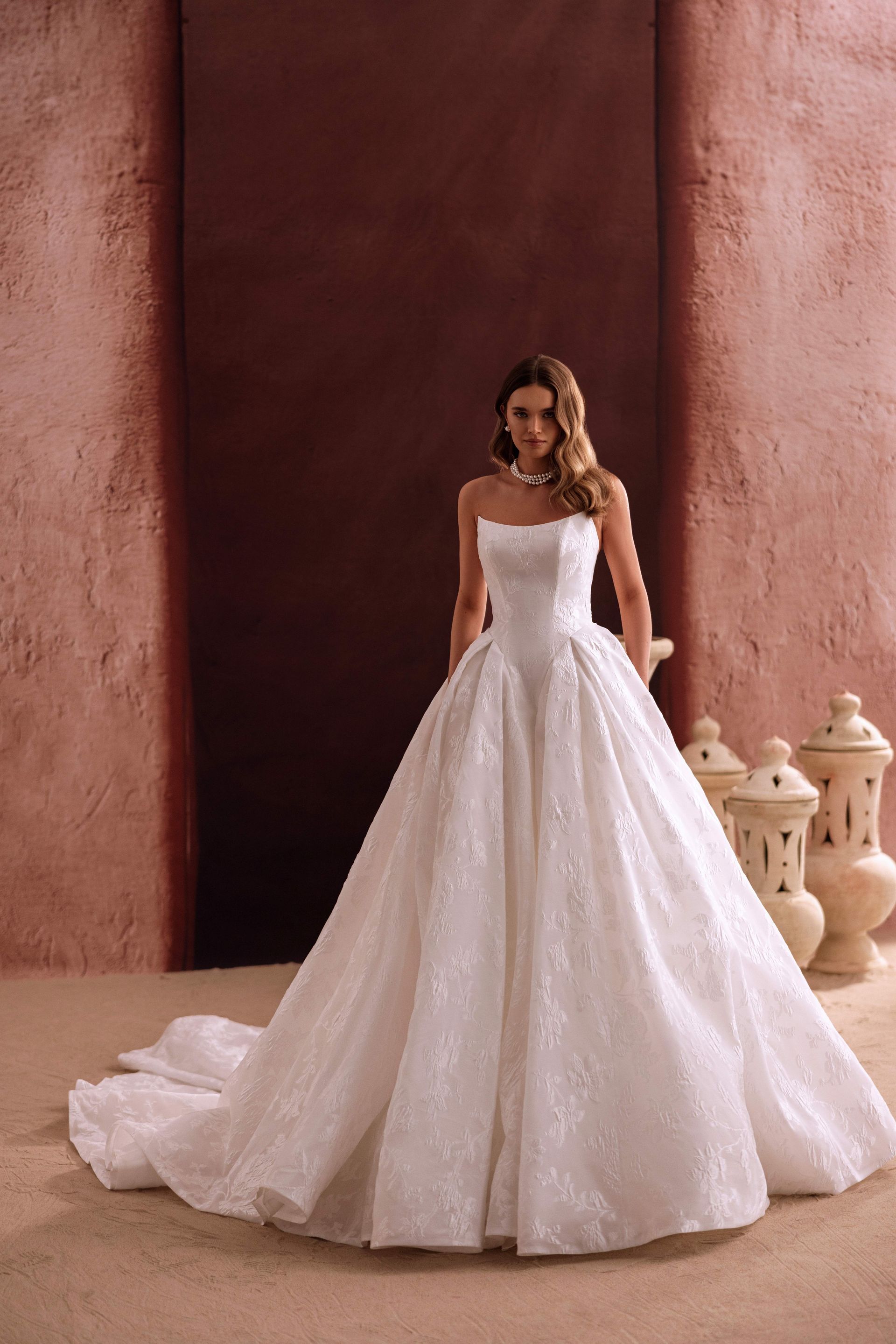 Frau im weißen trägerlosen Brautkleid, stehend, lächelnd, mit langer Schleppe, vor Terrakotta-Hintergrund.