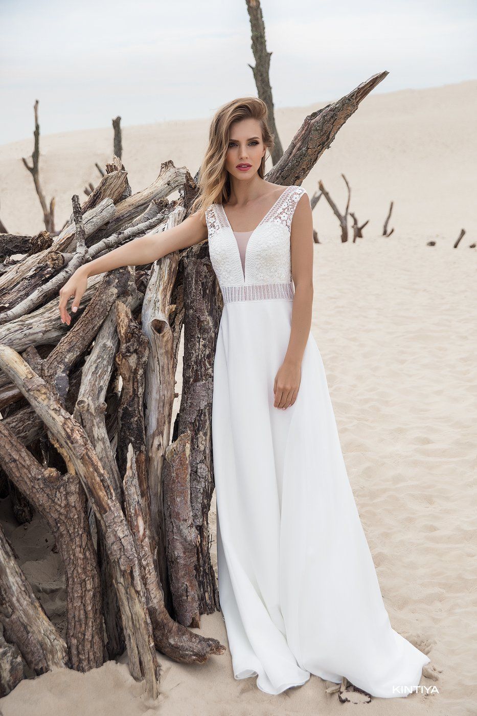 Eine Frau in einem weißen Brautkleid steht neben einem Stapel Baumstämme am Strand.