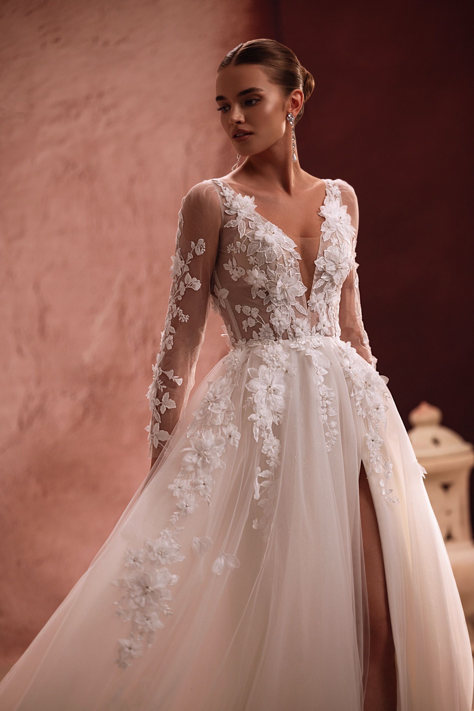 Frau im weißen Brautkleid mit floralen Details, langen Ärmeln und hohem Schlitz, posiert.