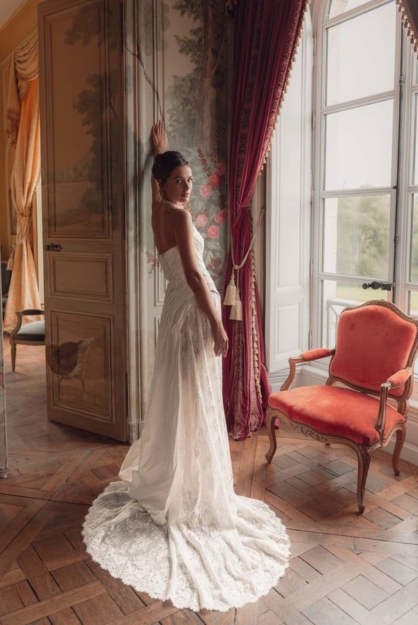 Eine Frau in einem weißen Brautkleid posiert neben einem Fenster in einem Raum mit floraler Wandkunst und roten Vorhängen.