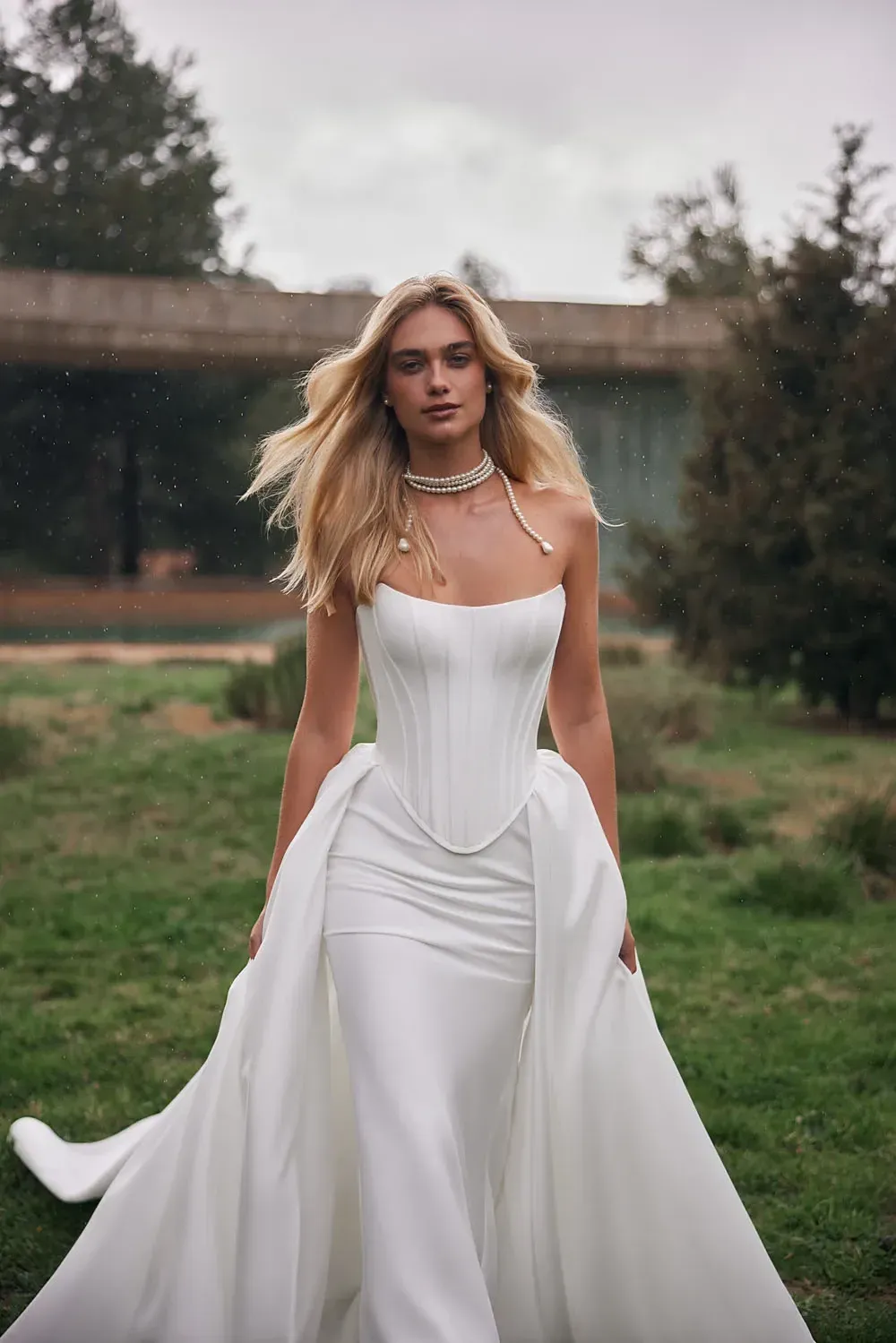 Frau im weißen Brautkleid geht draußen spazieren. Blondes Haar, Perlenkette, Wiese.