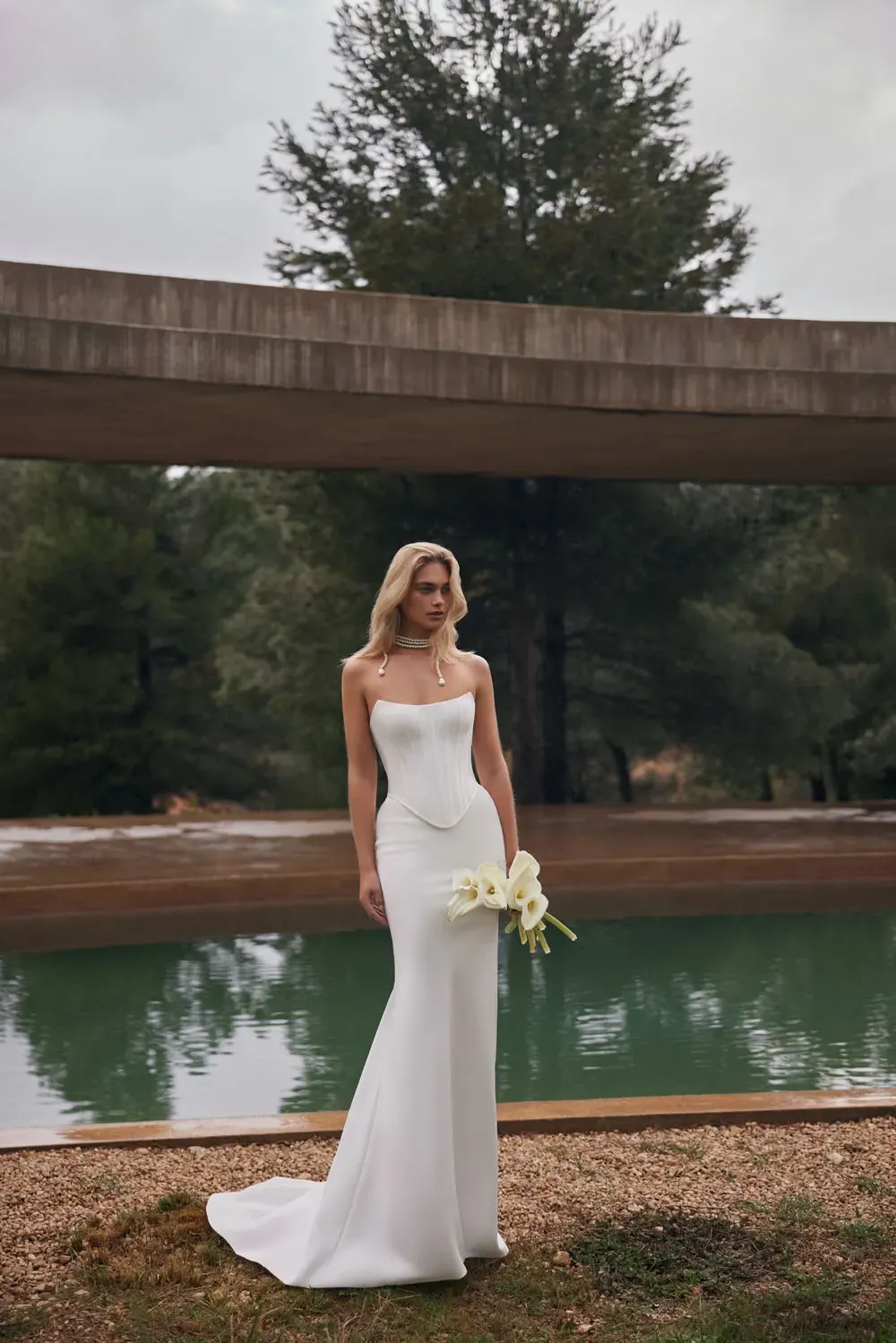 Blonde Frau im weißen Brautkleid mit Blumen in der Hand, steht neben einem Pool.