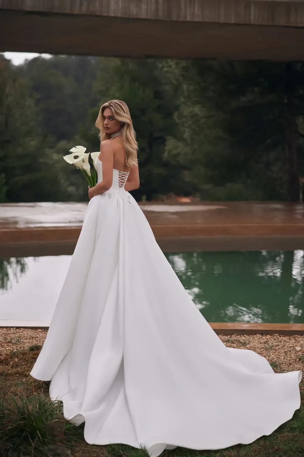 Braut in einem weißen trägerlosen Kleid, hält Blumen, blickt über ihre Schulter und steht an einem Pool.