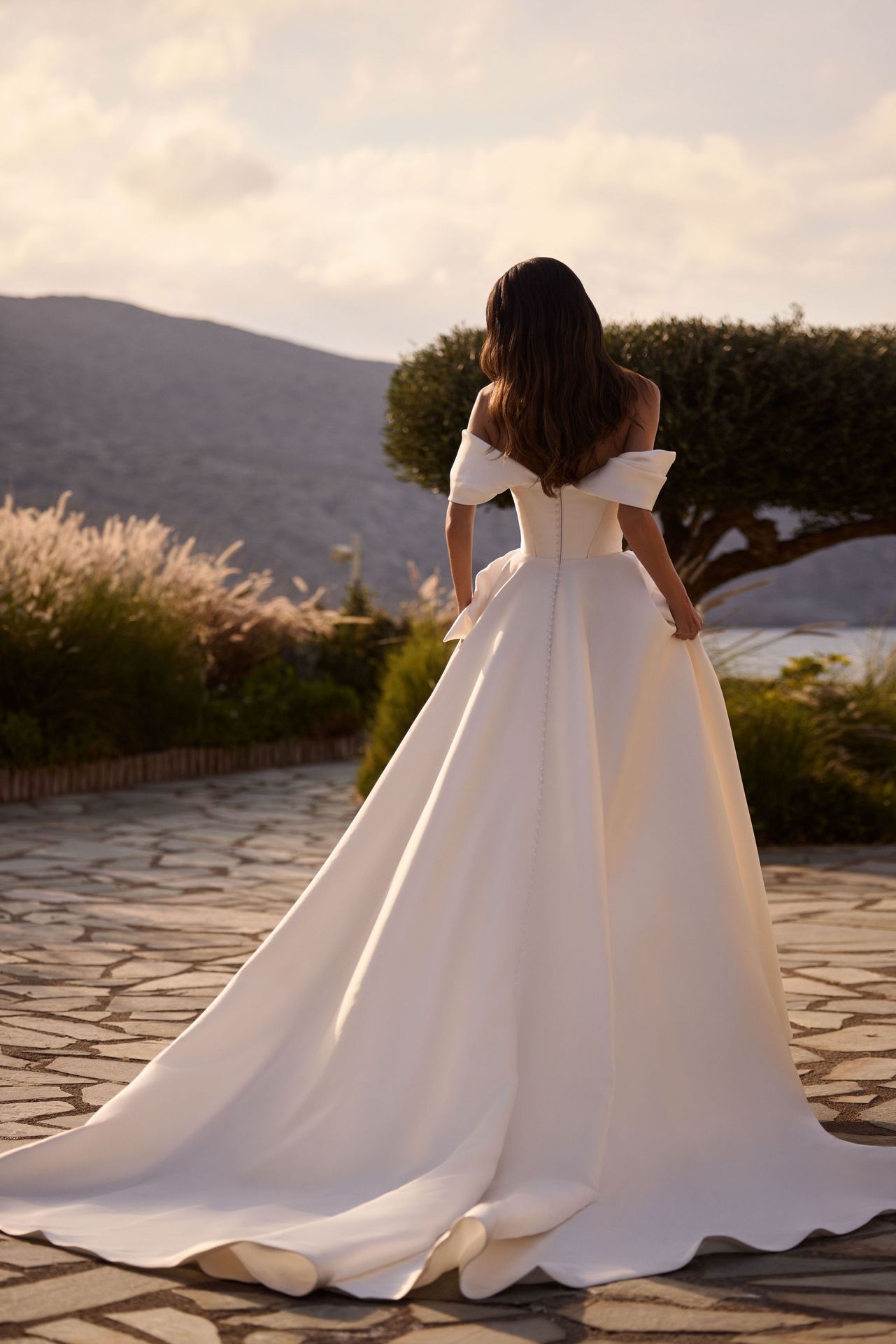 Frau im weißen schulterfreien Brautkleid, im Freien mit Bergen und Meer im Hintergrund.