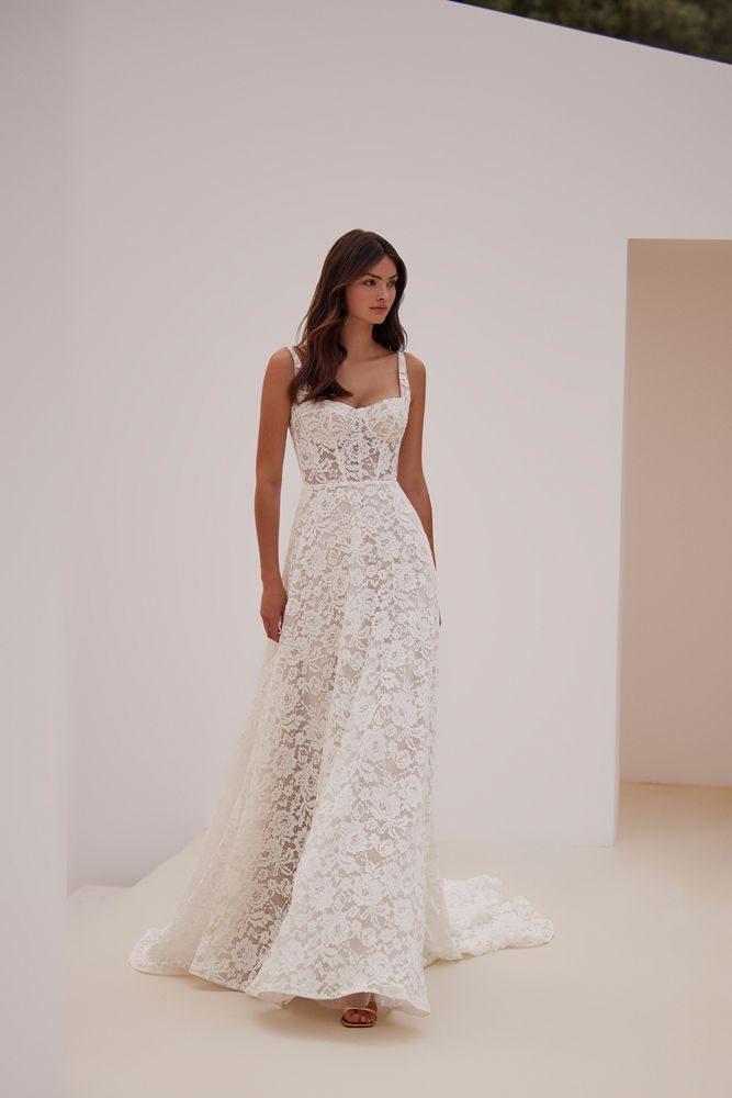 Eine Frau in einem weißen Spitzenbrautkleid steht vor einer weißen Wand.