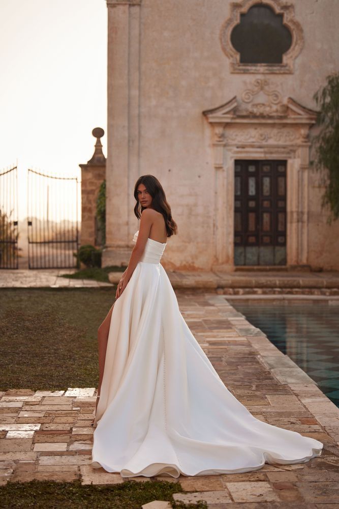 Frau in weißem trägerlosem Brautkleid mit hohem Schlitz steht in der Nähe eines Gebäudes und eines Pools.