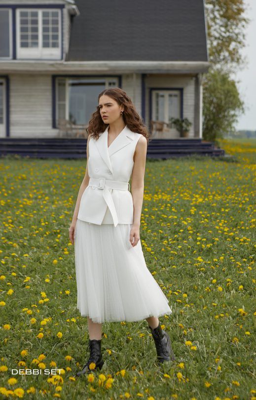 Eine Frau in einem weißen Kleid steht in einem Löwenzahnfeld vor einem Haus.