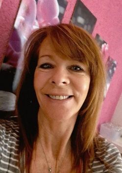 Une femme aux cheveux roux sourit, vêtue d'un haut bleu à fleurs, devant une œuvre d'art représentant une orchidée rose.