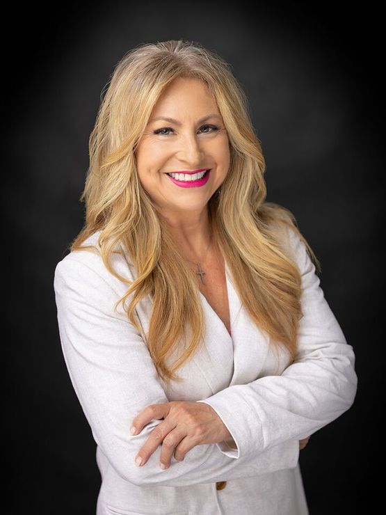 Smiling blonde woman in a white blazer with crossed arms against a dark background