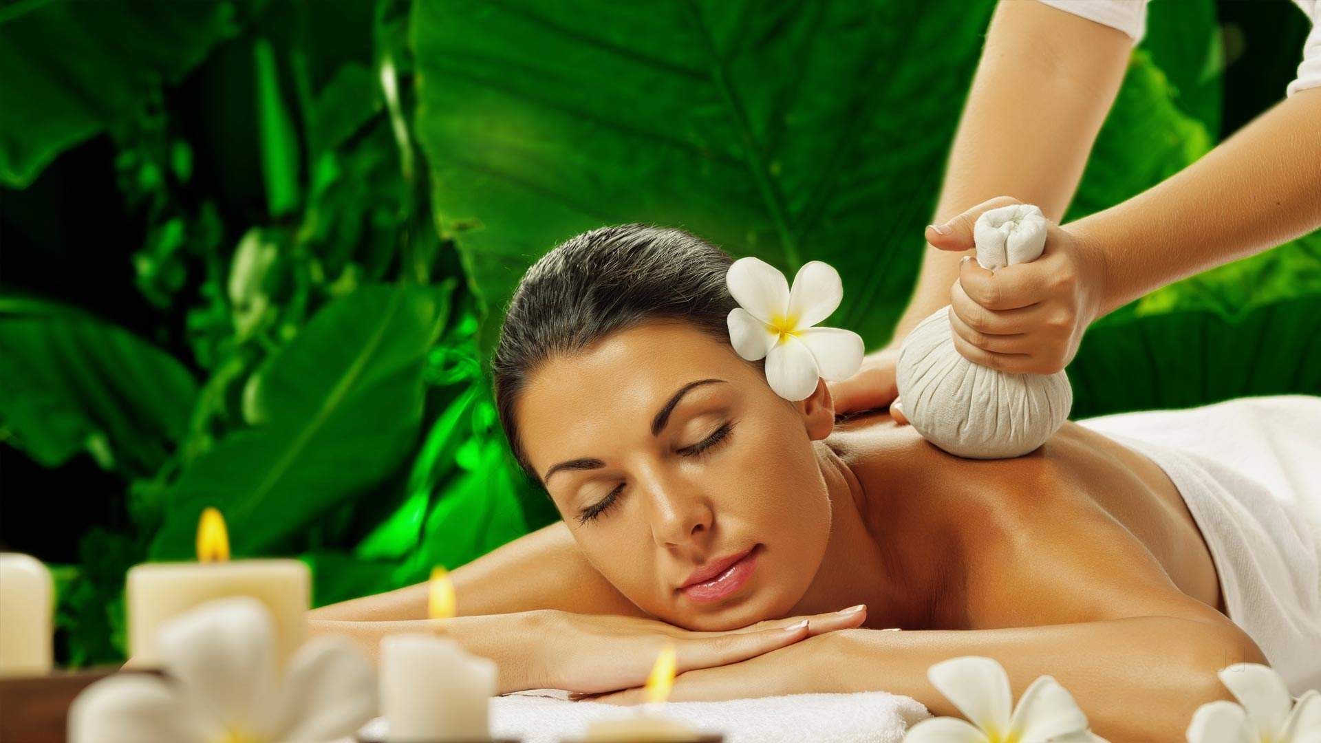 Woman receiving a spa treatment with herbal compresses on her back; lit candles and flowers in the foreground, lush green foliage background.